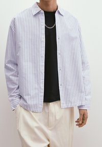 Chemise boutonnée rayée bleu clair et blanc, en coton, manches longues, coupe décontractée, portée sur un t-shirt noir, avec un détail de chaîne.