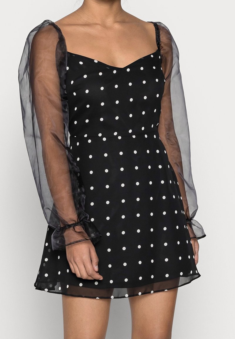 Czarna sukienka w białe kropki z dopasowanym górnym fragmentem, przezroczystymi bufkami i falbanami przy mankietach. Materiał jest lekki o zwiewnej strukturze.