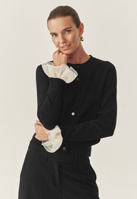 Crna kardigan s okruglim izrezom i gumbima, završena prozirnim bijelim volanima na rukavima. Uparen s odgovarajućim crnim hlačama.