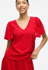 Sarkans V veida tops, izgatavots no gludas auduma ar puff piedurknēm un brīvu piegriezumu, ar divkāršu apakšmalu, kombinēts ar atbilstošām svārkiem.