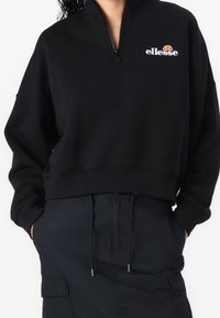 Musta collegepaita, valmistettu pehmeästä kankaasta, jossa on puolivetoketjullinen kaulus, lasketut olkapäät ja kirjailtu logo rinnassa.