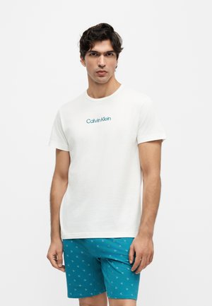 Mand iført hvid Calvin Klein t-shirt og blå shorts med lille gentagende logomønster, står mod hvid baggrund.