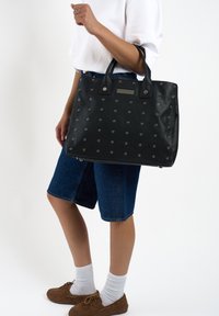 Zwarte leren tote bag met zilveren oogjes en een textuuraccent op de voorkant. Beschikt over dubbele handvatten en een gestructureerd ontwerp.