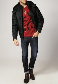 Chaqueta acolchada negra con capucha, usada sobre una camiseta gráfica roja. Vaqueros azul oscuro y botines marrones completan el conjunto.
