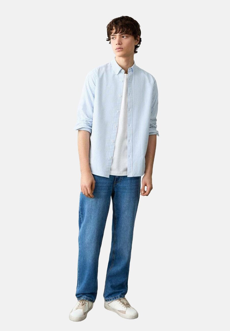Chemise à rayures bleu clair à manches longues portée sur un t-shirt blanc, associée à un jean bleu et des baskets blanches. Le mannequin est debout devant un fond uni.