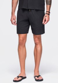 Homme portant un short noir décontracté et des sandales noires en tongs, debout les bras détendus le long du corps sur un fond uni.