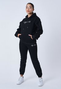 Sudadera con capucha negra y cordones, con bolsillo frontal y logo blanco, combinada con joggers negros ajustados que presentan el mismo logo. Zapatillas blancas.
