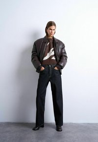 Femme portant une doudoune en cuir marron foncé, un pull à motifs marron, un jean noir à jambes larges et des bottes à talons noires, debout les mains dans les poches.