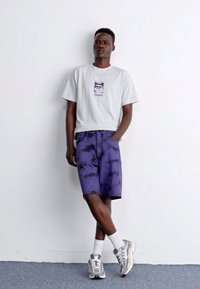 Camiseta gris de manga corta con un gráfico en el pecho, combinada con pantalones cortos de teñido anudado en púrpura. El modelo lleva zapatillas grises y calcetines blancos.