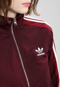 Bordeauxrode ritsjas met wit drie-strepen ontwerp op de schouders. Voorzien van een geborduurd wit Adidas-logo op de linkerborst. Gladde textuur.