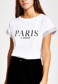 Bílá krátká trička s krátkým rukávem se nápisem "PARIS L'amour" černým písmem, s stříbrným zdobením na rukávech, v kombinaci s černými kalhotami.