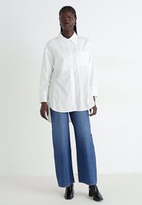 Camicia bianca a maniche lunghe con sottili righe verticali, colletto a bottoni, taschino sul petto; abbinata a jeans in denim blu a gamba larga e stivali neri.