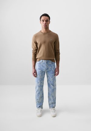 Suéter de punto marrón con mangas largas, jeans de camuflaje azul claro y zapatillas blancas. El suéter cuenta con un pequeño logo bordado en el pecho.