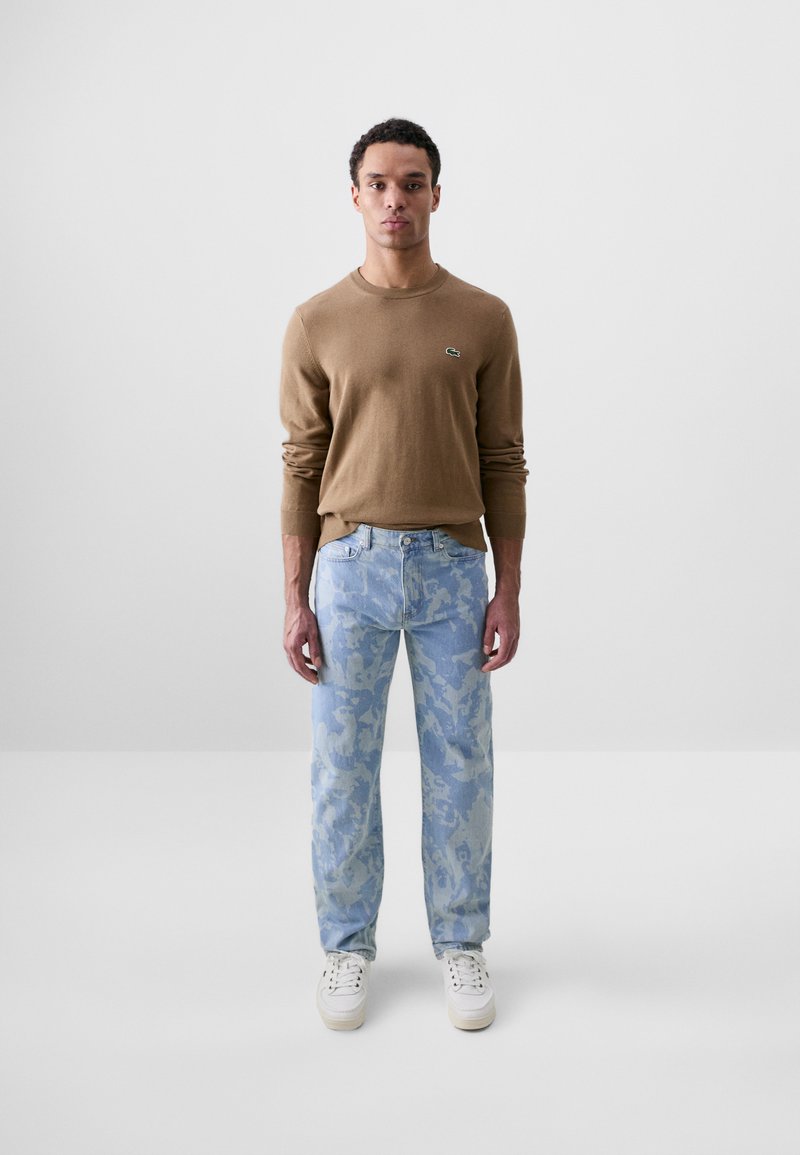 Suéter de punto marrón, jeans de camuflaje azul y zapatillas blancas. Diseño minimalista con un logo en el suéter.