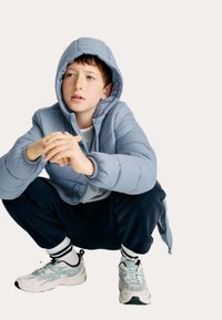 Pojke med fräknar som bär en ljusblå luvtröja, marinblå byxor och sneakers, hukar med händerna ihopknäppta och ser svagt åt sidan.