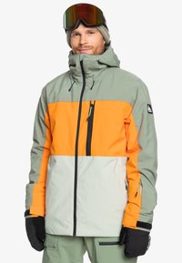 Chaqueta de esquí en verde y naranja, con cremallera frontal, cuello alto y puños ajustables. Material texturizado e impermeable con bolsillos con cremallera.