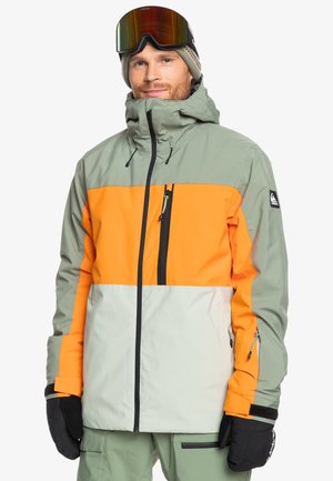 Chaqueta de esquí en verde y naranja, con cremallera frontal, cuello alto y puños ajustables. Material texturizado e impermeable con bolsillos con cremallera.