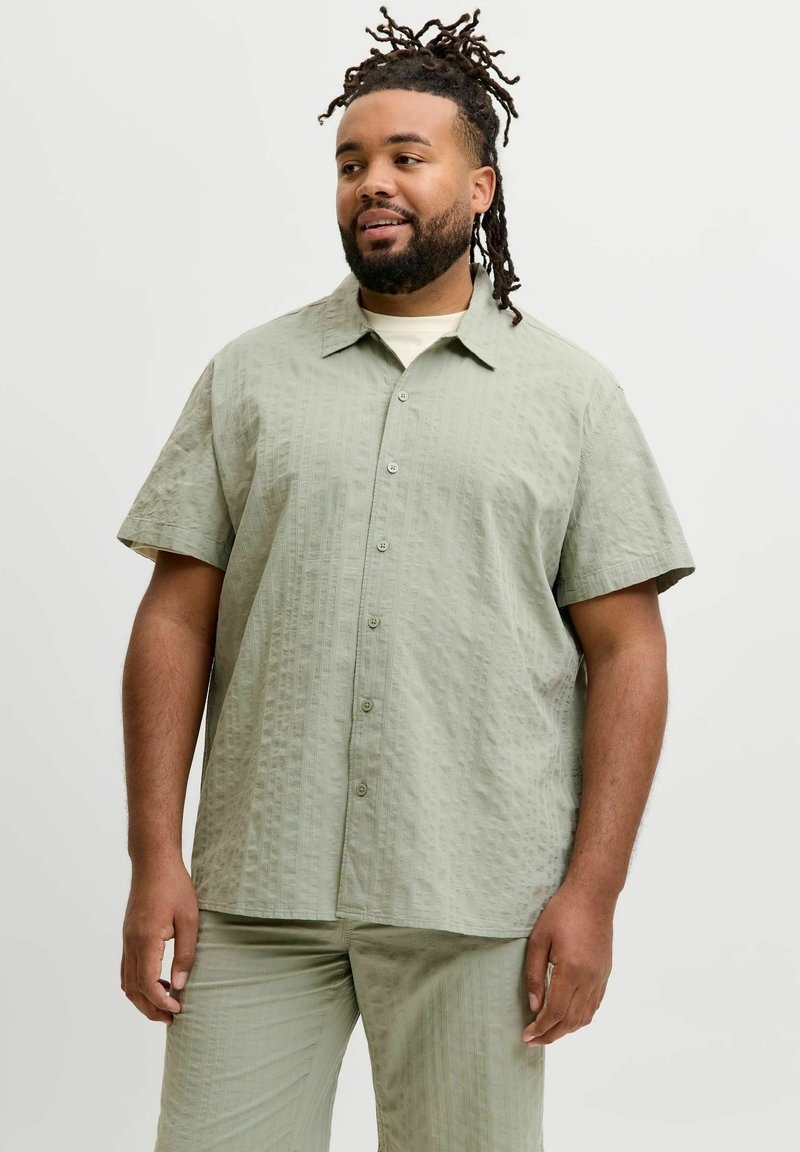 Άνδρας με μεσαίο τόνο δέρματος και dreadlocks, που φοράει ανοιχτό πράσινο πουκάμισο με κοντά μανίκια και κουμπιά και ταιριαστό παντελόνι, στέκεται μπροστά σε απλό λευκό φόντο.