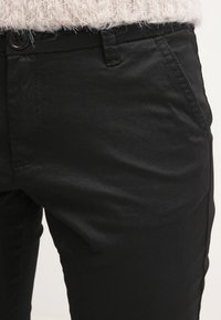 Pantalon noir en tissu lisse, avec poches avant et fermeture à bouton. Le design est ajusté avec une coupe droite.