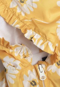 Giacca gialla a fiori con bordo arricciato. Il tessuto è leggero e liscio. Presenta una cerniera bianca e un motivo floreale in un delicato giallo e bianco.