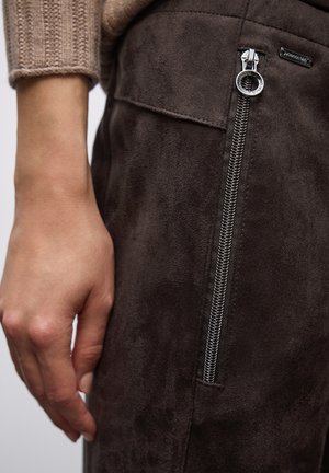Pantalones de ante marrón con un bolsillo lateral con cremallera, que cuenta con una cremallera plateada y una textura suave. Mano posicionada cerca de la cremallera para referencia de tamaño.