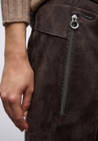 Brązowe zamszowe spodnie z boczną kieszenią na zamek, z metalowym zamkiem błyskawicznym i gładką fakturą. Ręka umieszczona blisko zamka dla skali.