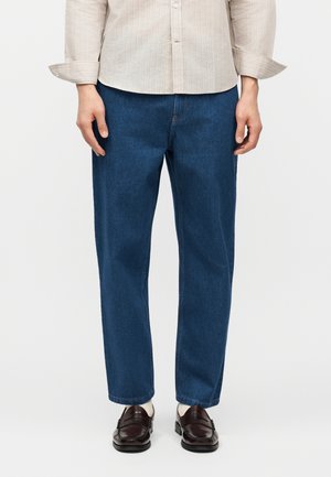 Άνδρας με τζιν παντελόνι κανονικής γραμμής, λευκό πουκάμισο με μακριά μανίκια, λευκές κάλτσες και σκούρα καφέ δερμάτινα μοκασίνια, στέκεται μπροστά σε απλό φόντο.