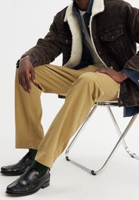 Brun kordflädesjacka med vit fårskinnskrage, kombinerad med kaki byxor och svarta lädermockasiner med gröna strumpor. Kromstol.