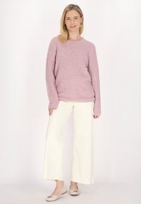 Rosa flauschiger Pullover mit langen Ärmeln, kombiniert mit cremefarbenen weit geschnittenen Hosen und beigen Ballerinas mit einem dekorativen Akzent. Heller Hintergrund.