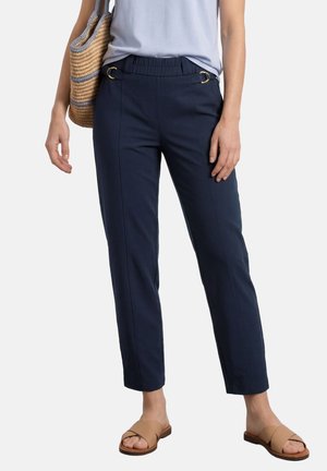 Kvinde iført mørkeblå ankellange bukser med guldbæltedetaljer, lyseblå top, brune slip-on sandaler og bærer en flettet taske på skulderen.