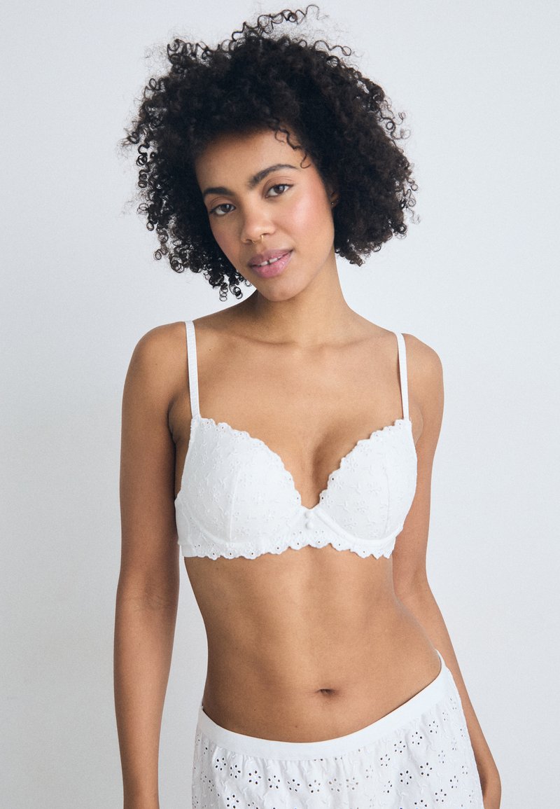Soutien-gorge en dentelle blanche avec des bords festonnés, présentant des motifs floraux et des bretelles fines ajustables. Tissu léger, conçu pour le confort et l'ajustement.