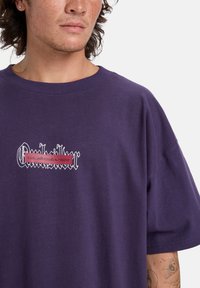 Lilla bomulds t-shirt med et grafisk logo i rød og hvid på brystet. Korte ærmer med en afslappet pasform og rund halsudskæring.