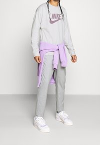 Šedá dlouhý rukáv Nike mikina s vínovým logem, světle šedé kalhoty a bílé tenisky s fialovými akcenty. Fialový svetr uvázaný kolem pasu.