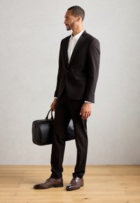 Svart dress med satenglapp, hvit skjorte, brune lær sko, holder en svart lærveske. Tergulv og lys grå bakgrunn.
