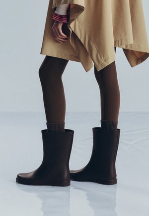 Person wearing dark brown tights, black mid-calf boots, and a beige asymmetric coat standing on a reflective surface.