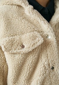 Beżowa, strukturalna kurtka z polaru z zatrzaskami noszona na czarnej koszuli z kołnierzykiem, pokazująca zbliżenie szczegółów materiału i zapięć.