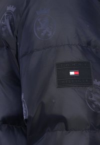 Marineblaue Steppjacke mit strukturiertem Stoff, dezentem Löwenwappenmuster und rechteckigem schwarzem Logopatch mit Markennamen und dreifarbigen Akzent.