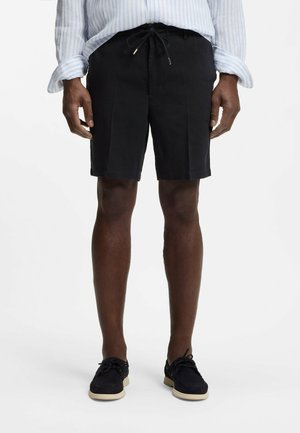 Mężczyzna w czarnych szortach ze sznurkiem, jasnoniebiesko-białej paskowanej koszuli z podwiniętymi rękawami oraz czarnych butach z białymi podeszwami.