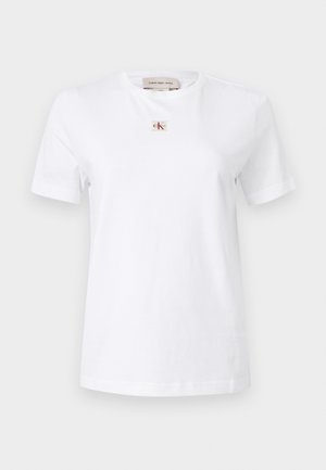 Tricou alb cu mânecă scurtă, având un mic patch cu logo-ul „CK” roșu și bej, centrat sub decolteu.