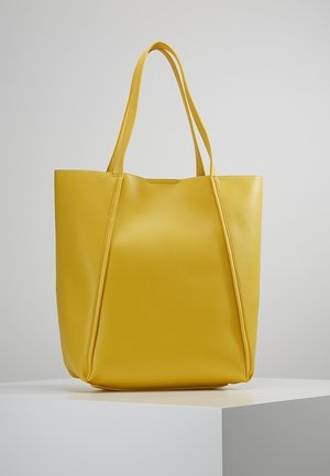 Żółta skórzana torba na ramię z dwoma paskami, stojąca pionowo na białym podeście na jednolitym szarym tle.