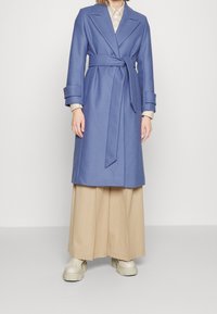 Abrigo azul de tela suave, con cinturilla, mangas largas y un cuello clásico. Combinado con pantalones anchos beige y zapatillas.