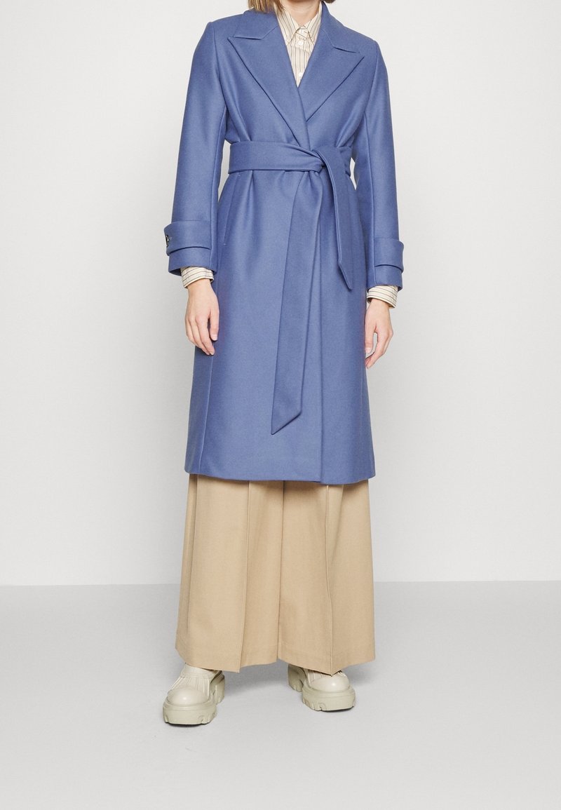 Abrigo azul de tela suave, con cinturilla, mangas largas y un cuello clásico. Combinado con pantalones anchos beige y zapatillas.