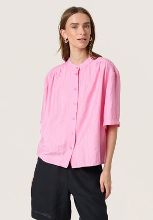 Vrouw draagt losse roze blouse met knopen en ellebooglange mouwen en zwarte korte broek, staand tegen een effen lichte achtergrond.