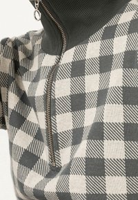 Haut zippé avec un col côtelé haut en vert foncé, associé à un motif à carreaux gris et beige. Détail de fermeture éclair en argent inclus.