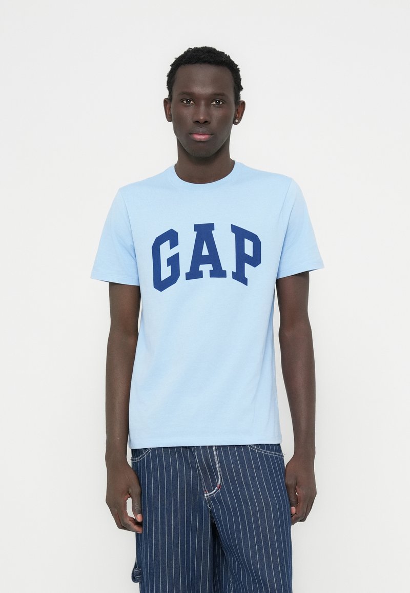 T-shirt en coton bleu clair avec un grand logo "GAP" bleu marine. Col rond et manches courtes, associé à un pantalon foncé à fines rayures.
