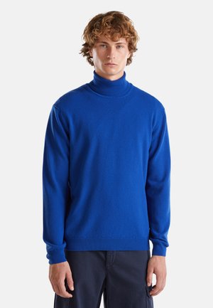 Jonge man met krullend haar, gekleed in een felblauwe coltrui en donkere broek, staande tegen een effen witte achtergrond.