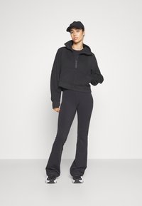 Haut en sweatshirt noir cropped avec col haut et zip à moitié ouvert, associé à un pantalon noir évasé. Une casquette noire et des chaussures de sport complètent le look.