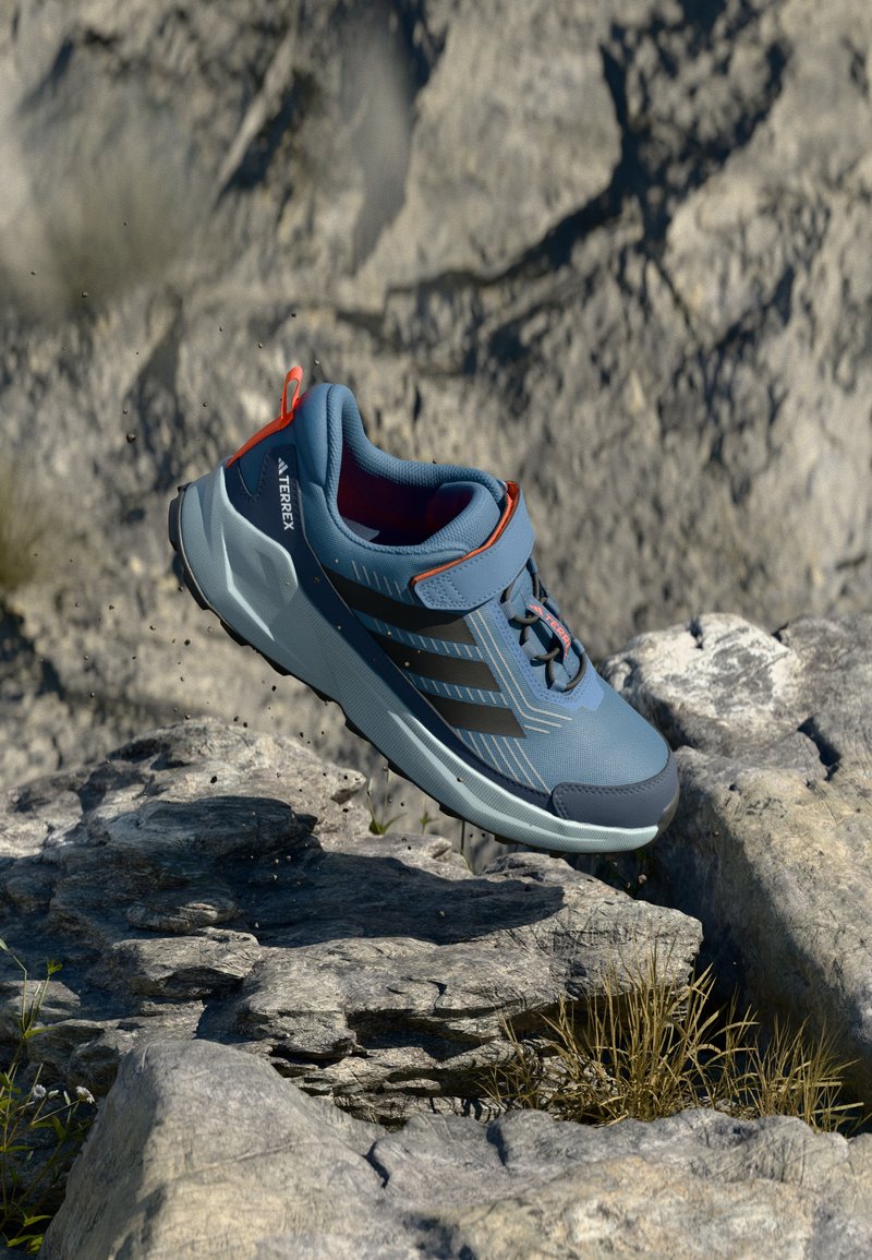 Blå idrettssko med glatt tekstur, med svarte striper, røde detaljer og en dempet såle, plassert midt i luften over steinete terreng.