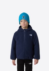 Granatowa polarowa kurtka z kapturem i przednim zamkiem błyskawicznym, w parze z jaskrawo niebieską prążkowaną czapką z naszywką z niedźwiedziem; czarne spodnie pod spodem.