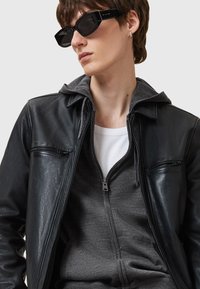 Veste en cuir noir avec poches zippées, sweat à capuche gris en dessous, et t-shirt blanc. Lunettes de soleil carrées noires. Texture de tissu doux.